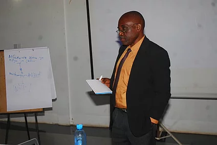 A man stands in a seminar room, holding a piece of paper as he presents. He wears a black suit with an orange shirt. To his left is a flip chart with handwritten text. In front of him is a table with a blue water bottle and electronic equipment. A projector screen is visible in the background, reinforcing the educational setting focused on research methods.