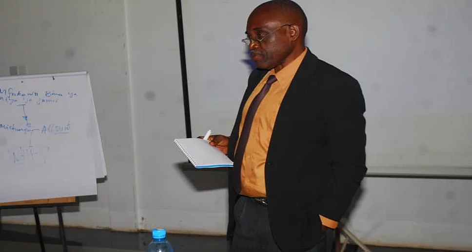 A man stands in a seminar room, holding a piece of paper as he presents. He wears a black suit with an orange shirt. To his left is a flip chart with handwritten text. In front of him is a table with a blue water bottle and electronic equipment. A projector screen is visible in the background, reinforcing the educational setting focused on research methods.
