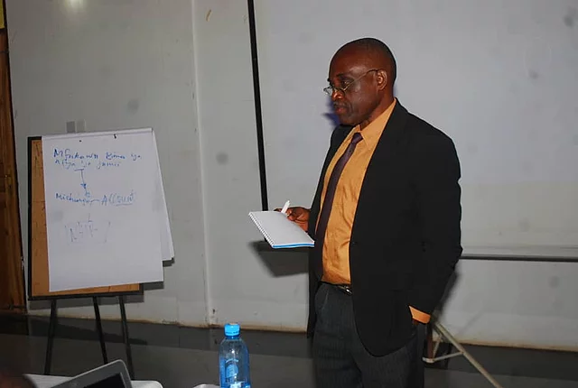 A man stands in a seminar room, holding a piece of paper as he presents. He wears a black suit with an orange shirt. To his left is a flip chart with handwritten text. In front of him is a table with a blue water bottle and electronic equipment. A projector screen is visible in the background, reinforcing the educational setting focused on research methods.