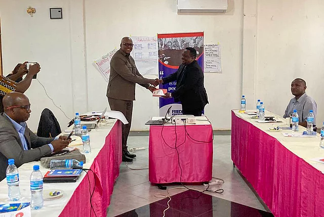 A formal workshop or meeting with participants seated around a U-shaped table arrangement. Two men stand in the center, shaking hands—possibly marking a presentation or agreement—while another person captures the moment with a photo. The tables are covered with white and pink cloths and display bottled water, notebooks, and documents. Behind the handshake, a banner reads “FRIEDRICH EBERT STIFTUNG” and “It starts with people,” indicating the involvement of the Friedrich Ebert Foundation. Posters with handwritten notes are visible on the wall, suggesting interactive group work or brainstorming