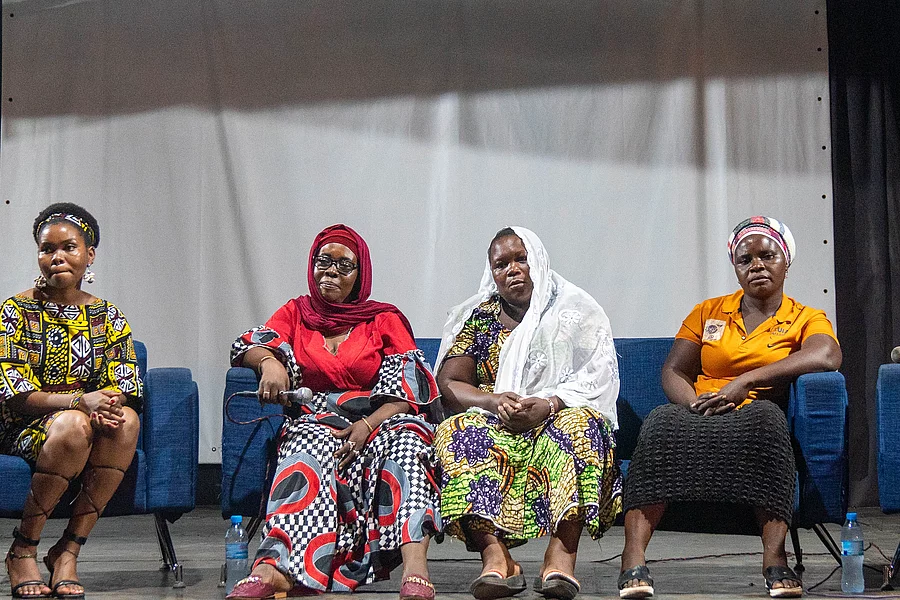 Five individuals seated on a stage during a panel discussion, each wearing vibrant, traditional African attire. They sit in a row on blue chairs, facing the audience. Behind them is a large white screen, suggesting a presentation or visual display. The indoor setting is well-lit, highlighting the cultural richness and formal tone of the event.
