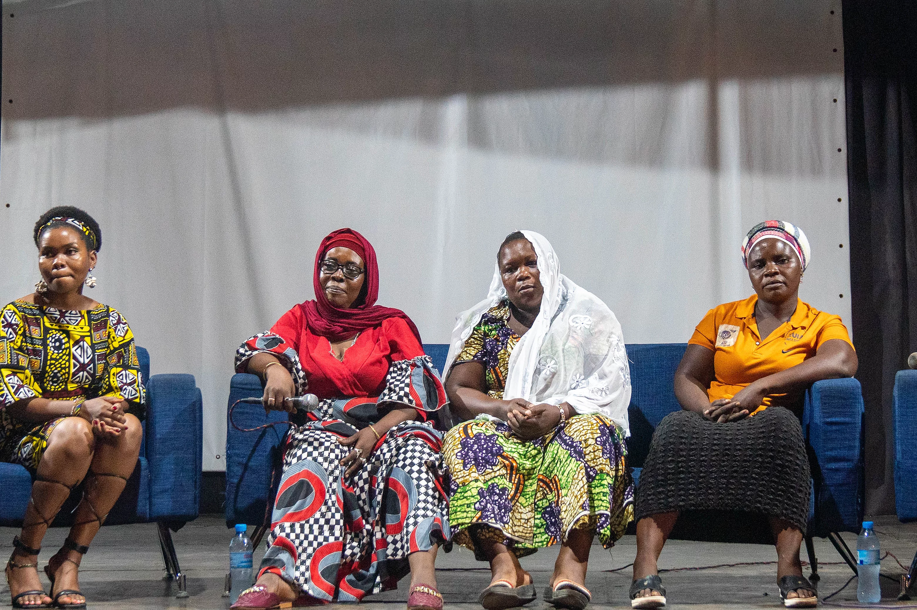 Five individuals seated on a stage during a panel discussion, each wearing vibrant, traditional African attire. They sit in a row on blue chairs, facing the audience. Behind them is a large white screen, suggesting a presentation or visual display. The indoor setting is well-lit, highlighting the cultural richness and formal tone of the event.