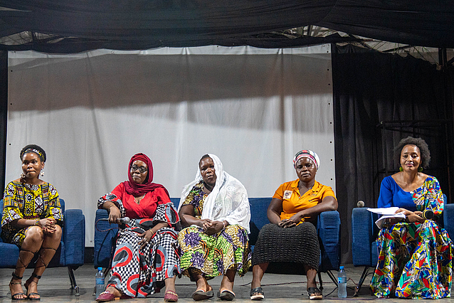 Five individuals seated on a stage during a panel discussion, each wearing vibrant, traditional African attire. They sit in a row on blue chairs, facing the audience. Behind them is a large white screen, suggesting a presentation or visual display. The indoor setting is well-lit, highlighting the cultural richness and formal tone of the event.