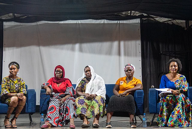 Five individuals seated on a stage during a panel discussion, each wearing vibrant, traditional African attire. They sit in a row on blue chairs, facing the audience. Behind them is a large white screen, suggesting a presentation or visual display. The indoor setting is well-lit, highlighting the cultural richness and formal tone of the event.