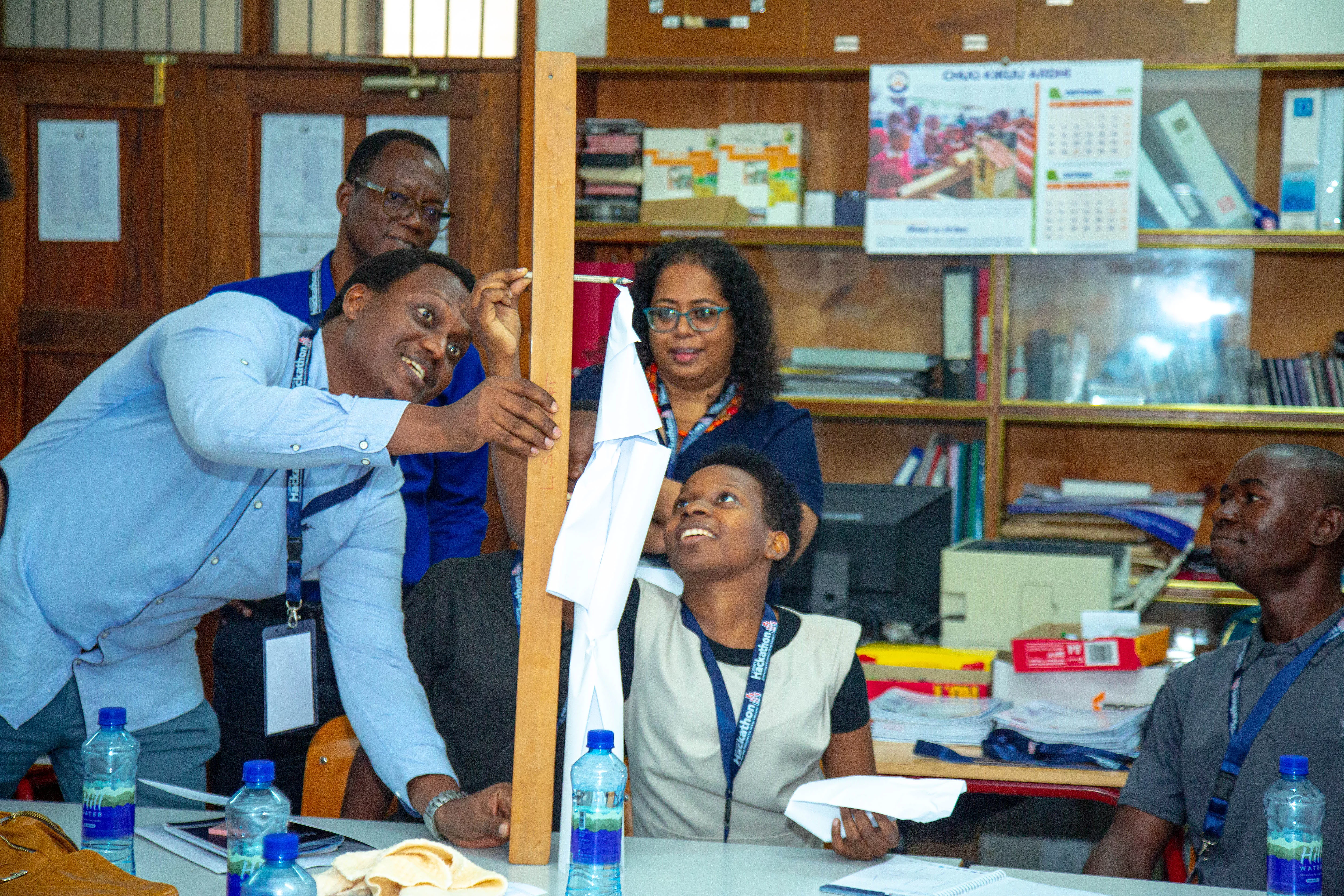 A group of adults participating in a hands-on educational activity in a classroom or office setting. One person holds a vertical wooden plank with cloth attached while others observe and engage. Participants wear lanyards, and the background includes shelves with books, files. Bottled water, notebooks, and materials on the table reinforce the collaborative learning environment.