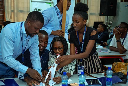 A group of individuals participate in a collaborative activity around a table, likely part of a hackathon event, as indicated by visible lanyards and name tags labeled “Hackathon.” They are actively engaged in constructing paper structures, with notebooks, water bottles, and other materials scattered across the table. The setting resembles a classroom or workshop, and other participants are visible in the background, observing or working. The scene captures teamwork, creativity, and problem-solving in an educational or innovation-driven environment.
